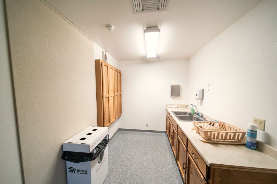 A plain office break room kitchenette with a double sink, wood cabinets, and a recycling box