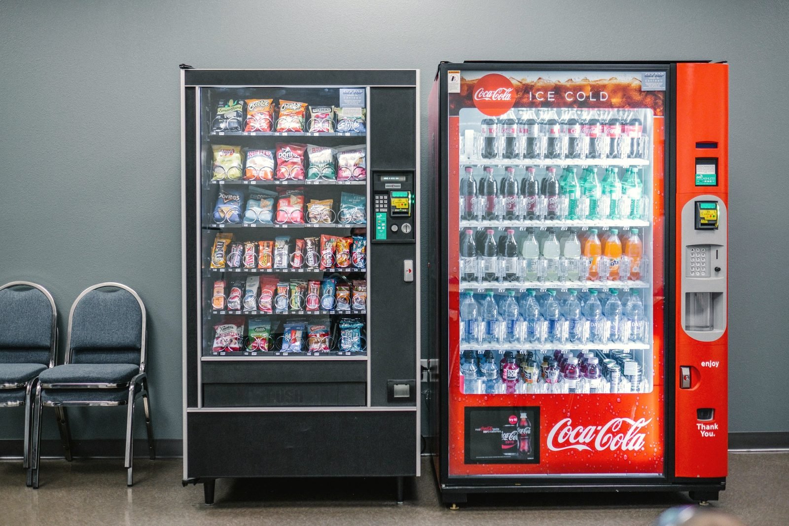 A snack and a cold-drink vending machine standing side by side against a gray office wall, waiting chairs beside them
