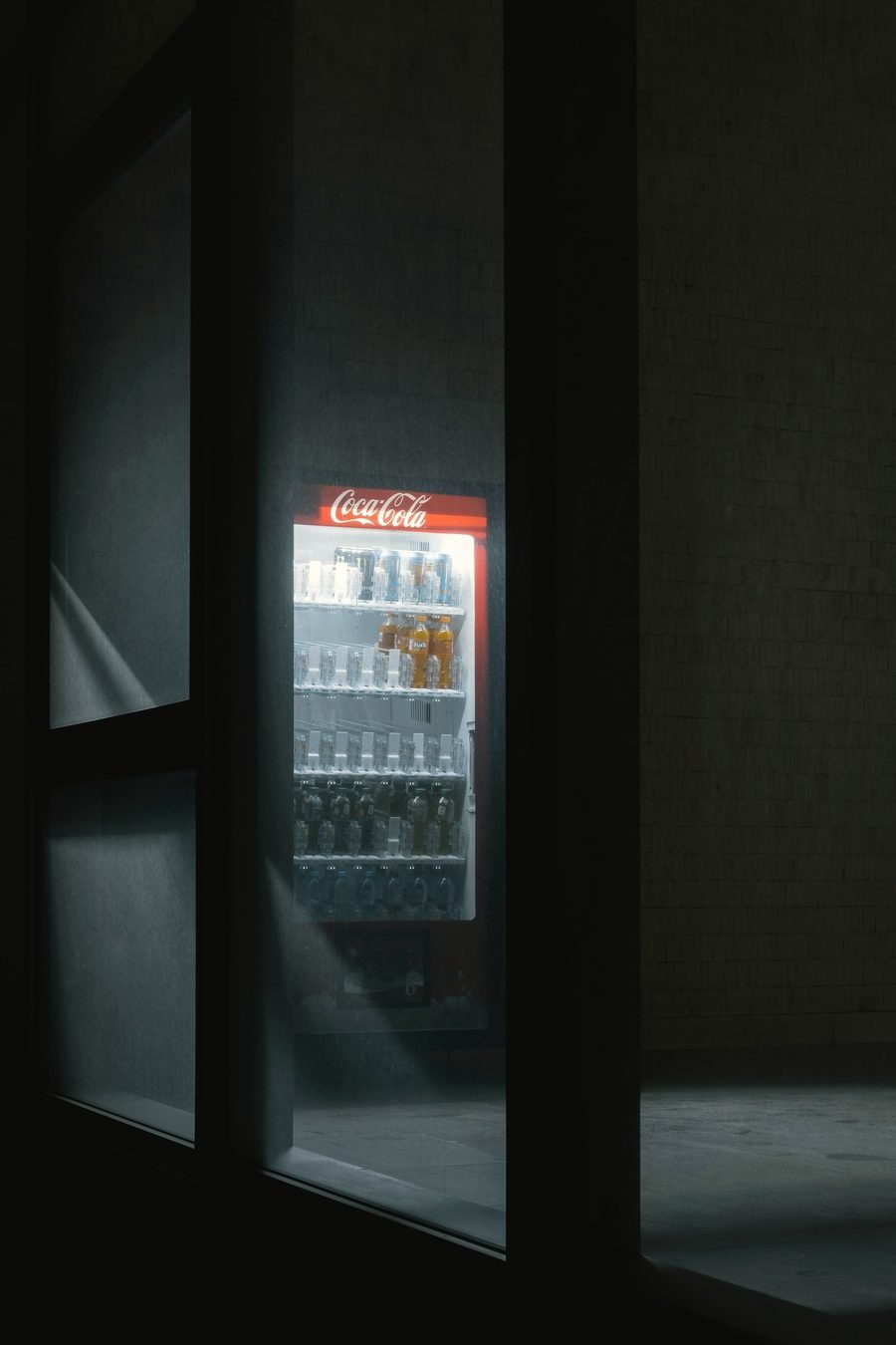 A lit Coca-Cola cooler glimpsed through a dark hallway at night