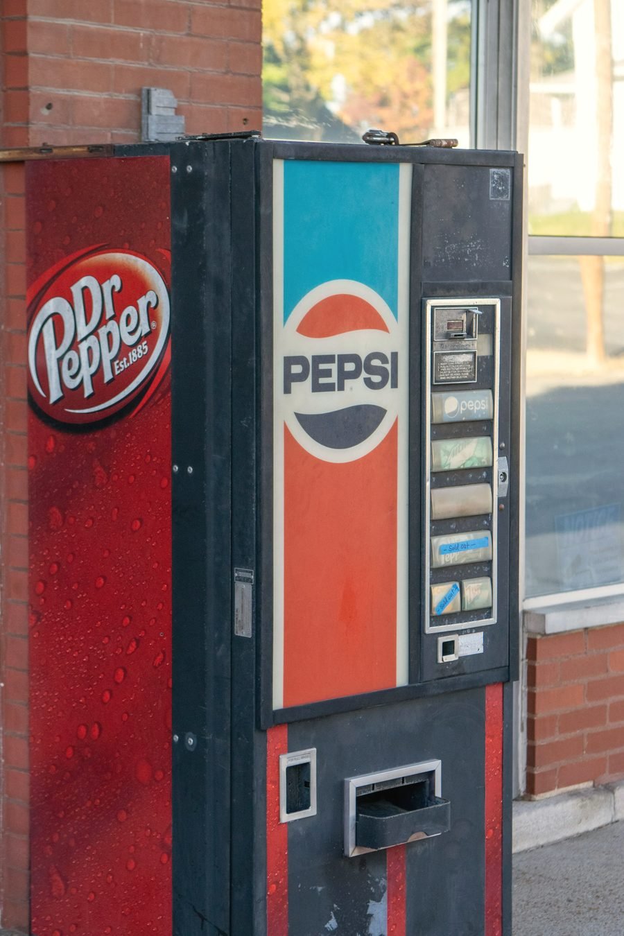A weathered Pepsi and Dr Pepper vending machine outside a brick storefront